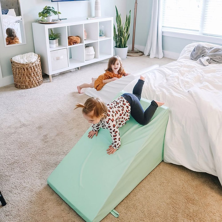 Child sliding down triangular foam wedge placed next to a bed