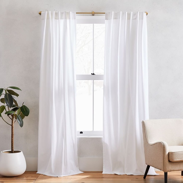 White floor length curtains next to small indoor plant and beige chair. 