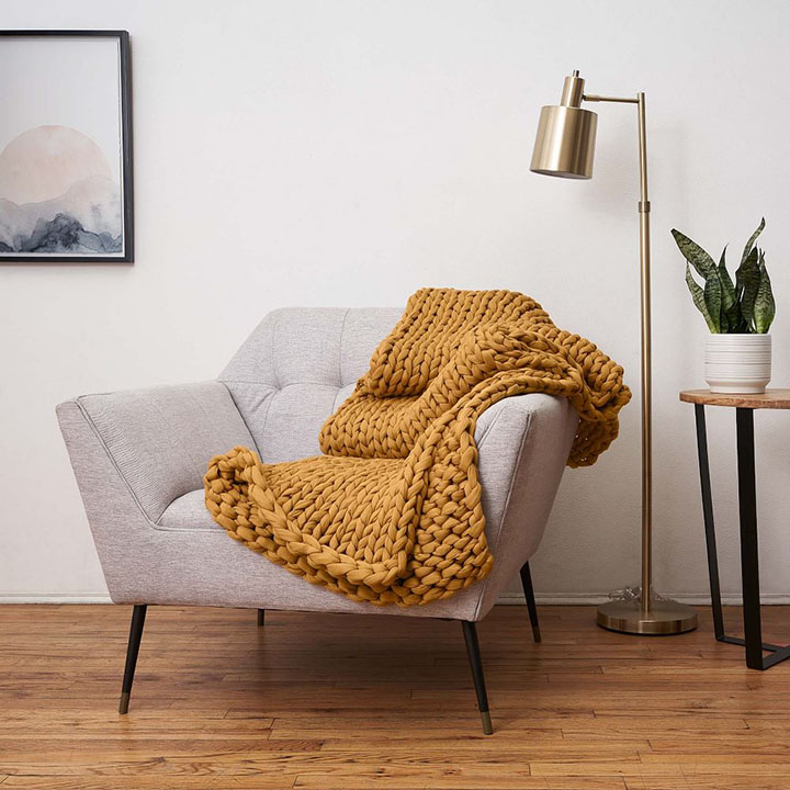 Camel chunky knit blanket on gray accent chair next to bronze floor lamp and wood side table with small plant.