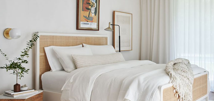 White and beige wicker bedframe with white bedding, wood nightstand and white area rug. 