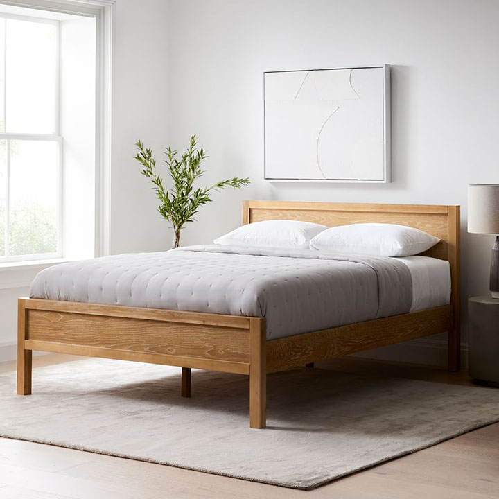 Natural wood bedroom, white and gray bedding, beige area rug, and large plant in corner. 