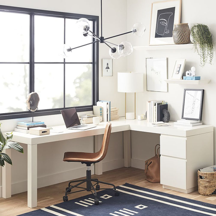 White l-shaped desk with bulb light fixture, picture frames and blue rug. 