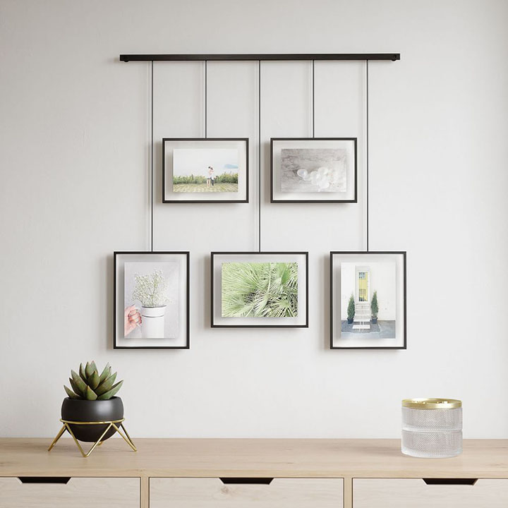 Five black picture frames creating a gallery wall with a wood dresser, small plant, and candle.