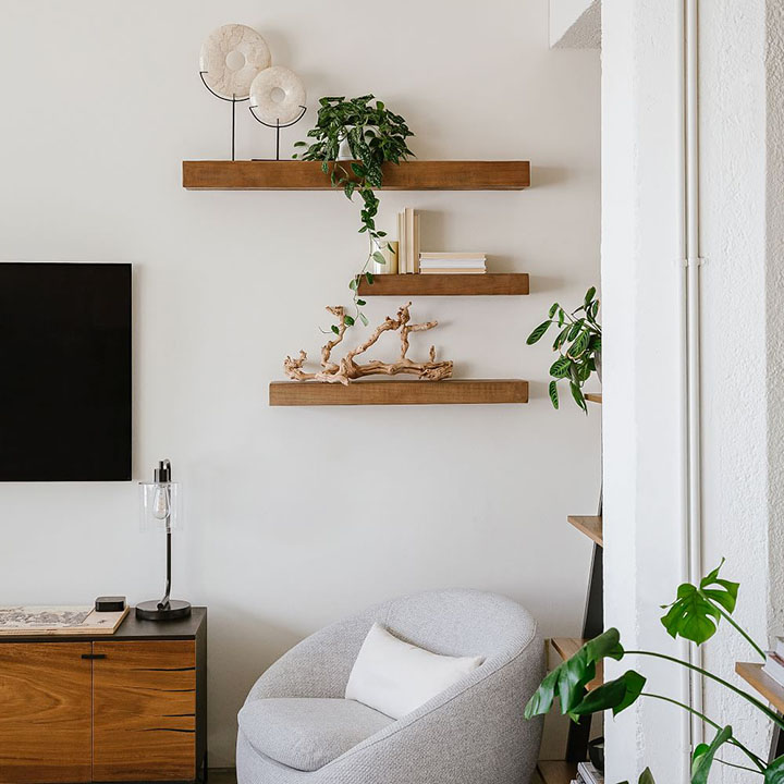 Three floating wood shelves above gray chair next to wood media console and TV. Three floating wood shelves above gray chair next to wood media console and TV.