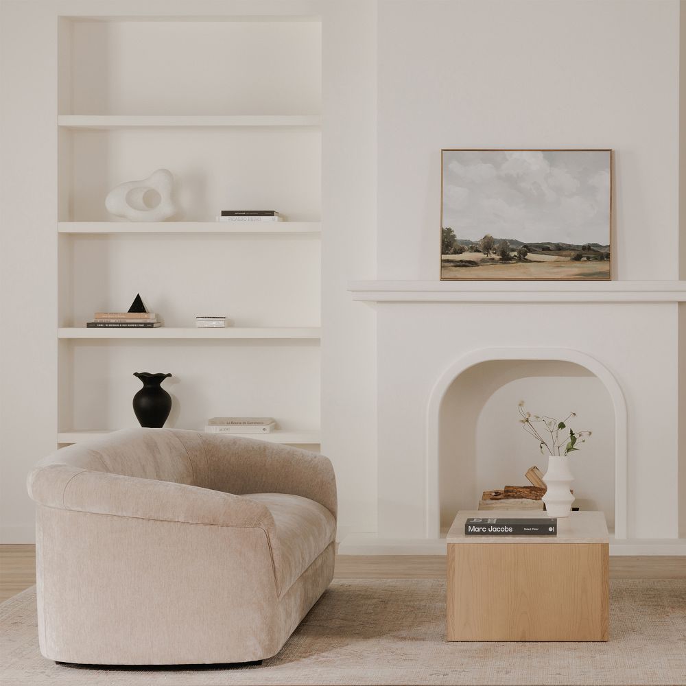 Neutral living room with a beige upholstered sofa, a wooden side table, and built-in white shelves beside a white fireplace; a framed landscape painting rests on the mantel.