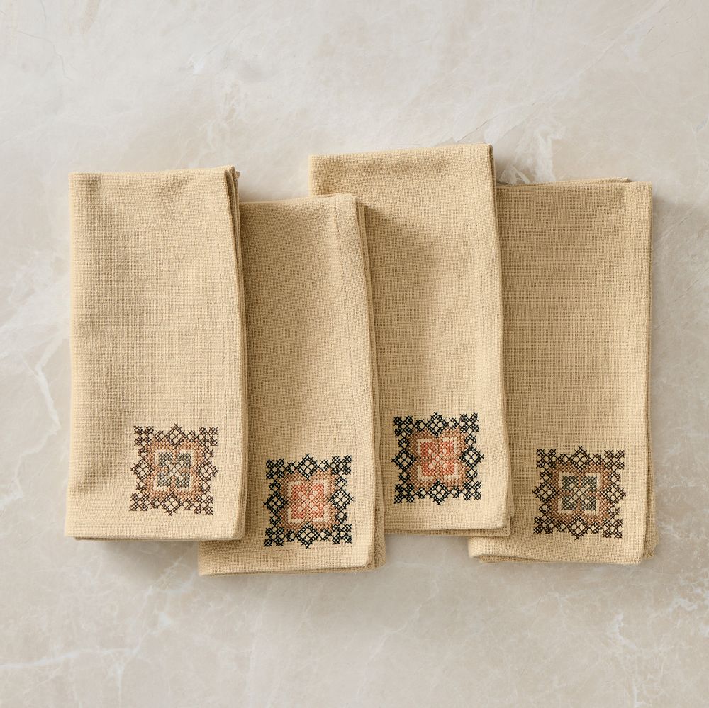 Four flax-colored napkins, each with a small stitched medallion in green and rust