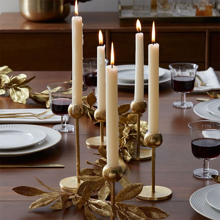 Long candles in brass candleholders in the center of a table with gold leaves. 