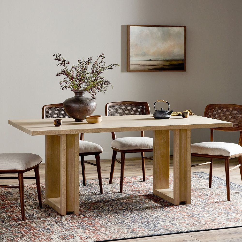 A wooden dining table with a rectangular tabletop and thick sturdy legs, surrounded by chairs on a patterned rug in a dining room.