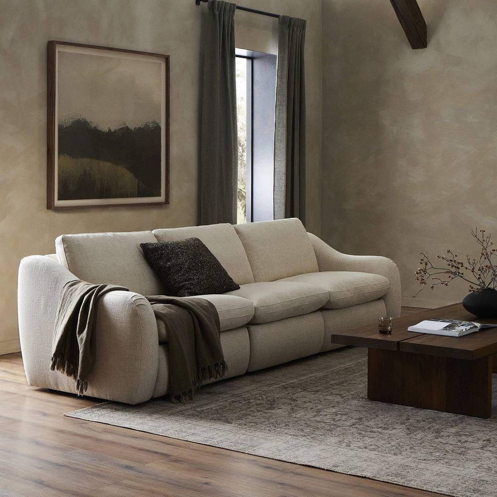 A beige fabric sofa with rounded arms in a living room setting, a dark brown throw blanket draped over the left arm, and a black textured throw pillow.