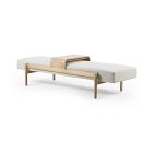 A light wood frame daybed with white mattress and a pale blanket, small wooden shelf behind